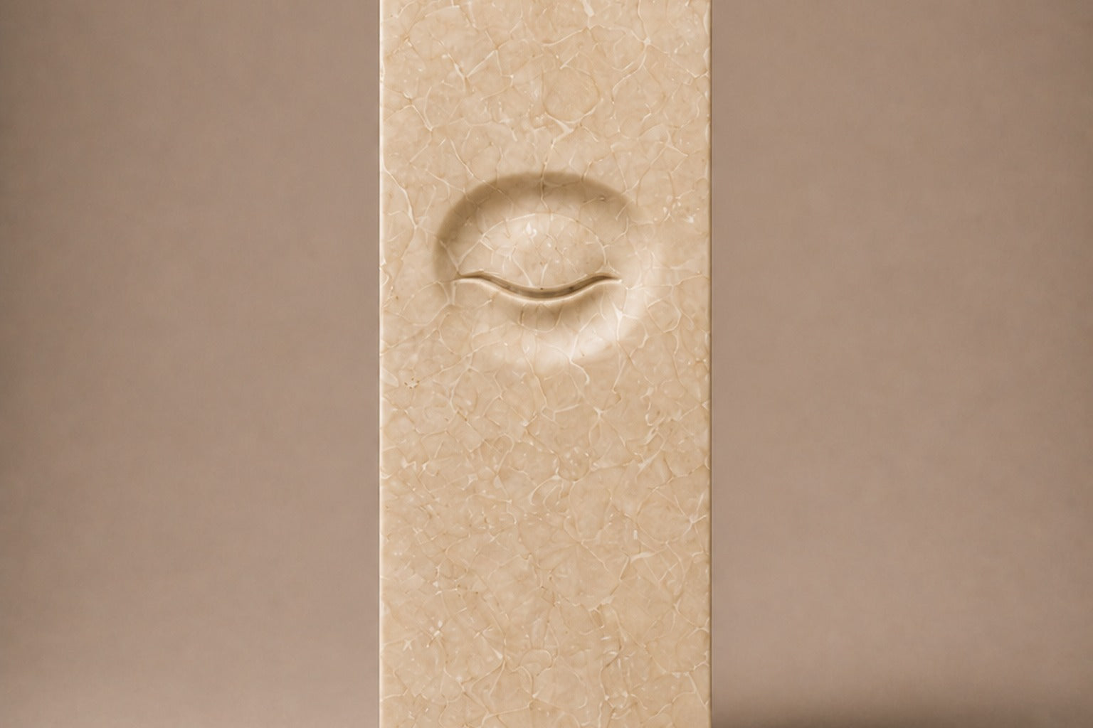 Tall cream Crema Marfil marble pedestal with a closed eye carved in shallow relief on one face.