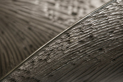 Wide woven-wire infinity form in pale silver-grey with two lens voids, against a plaster wall.