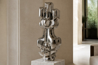 Mirror-polished stainless steel sculpture Alkaid, a column of stacked interlocking blocks, on a white marble cube plinth.
