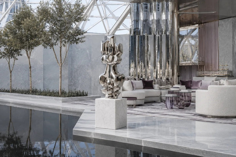Mirror-polished stainless steel sculpture Alkaid, a column of stacked interlocking blocks, on a white marble cube plinth.