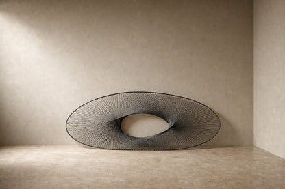 Broad woven-wire ellipse in pale grey with an open oval void, against a plaster wall.