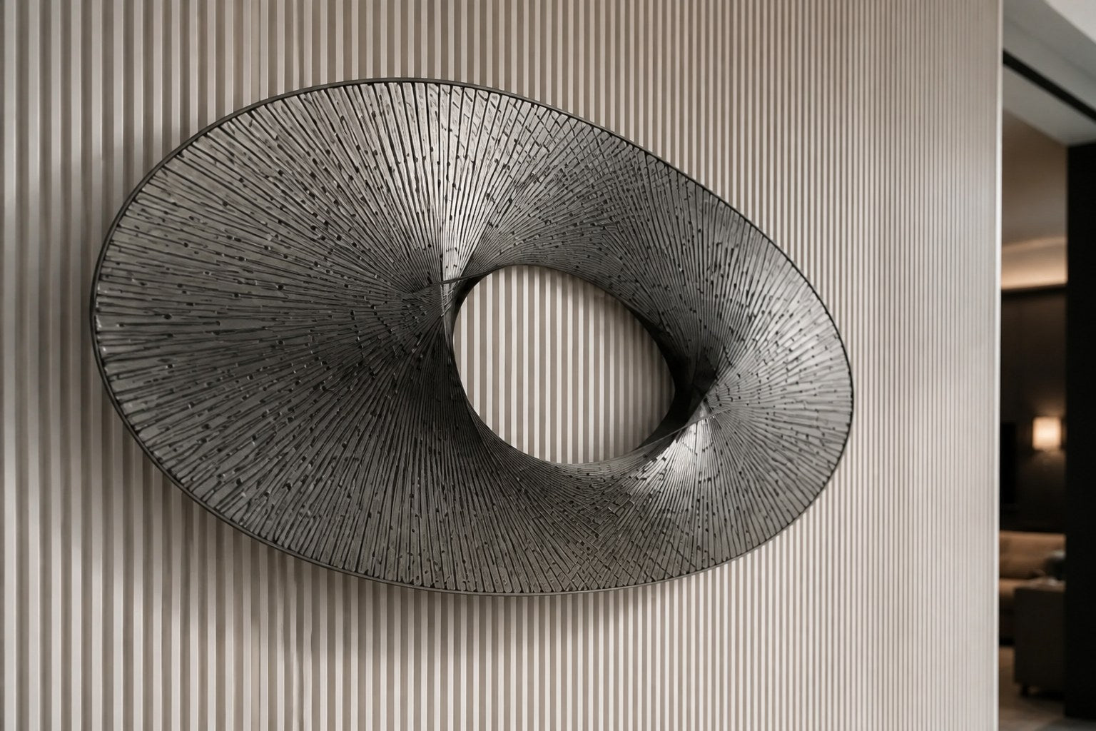 Broad woven-wire ellipse in pale grey with an open oval void, against a plaster wall.