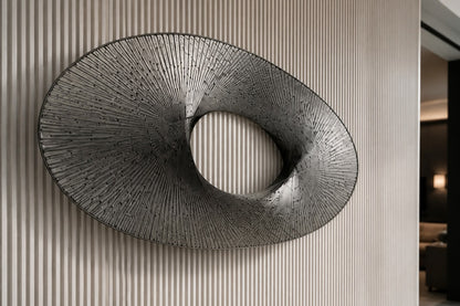 Broad woven-wire ellipse in pale grey with an open oval void, against a plaster wall.