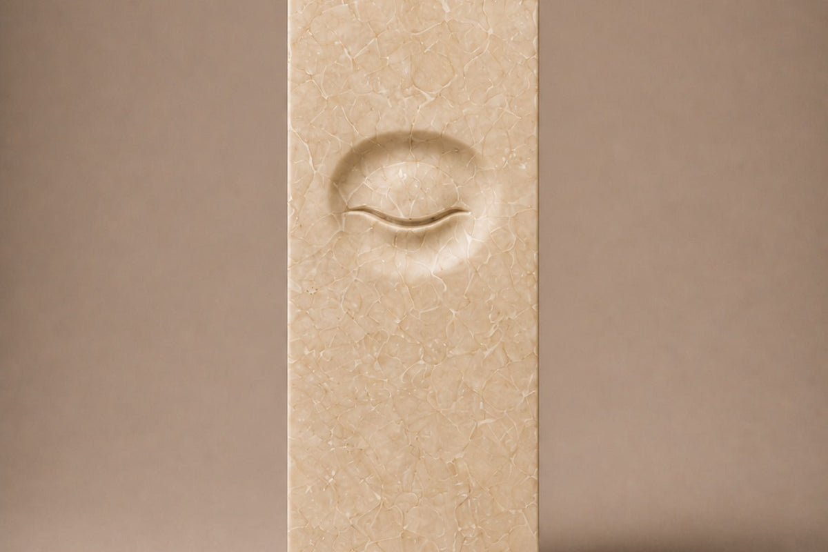 Tall cream Crema Marfil marble pedestal with a closed eye carved in shallow relief on one face — front elevation