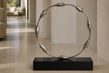 Mirror-polished stainless steel ring sculpture Estero Ø70, eight linked forms nose to tail, on a black granite block.