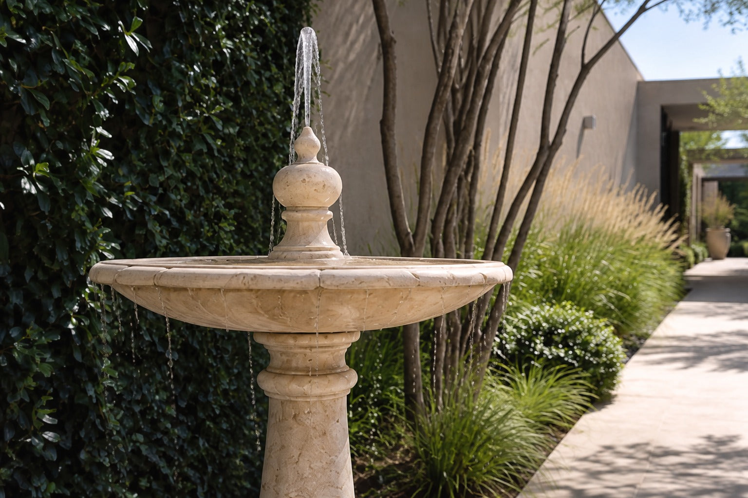 Pale limestone pedestal fountain Fontena Ø65, cream with open fossil speckle, on a pale stone floor.