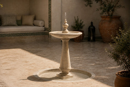 Pale limestone pedestal fountain Fontena Ø65, cream with open fossil speckle, on a pale stone floor.