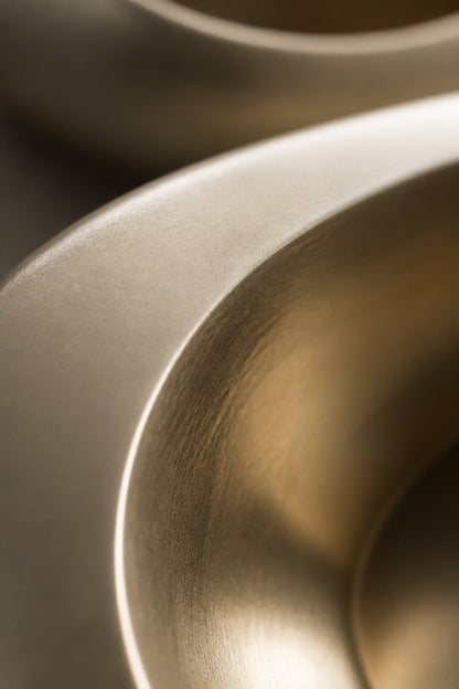 Champagne gold bronze wall sculpture Guijarro, a continuous folded ribbon swelling to a smooth belly, on a plaster wall.