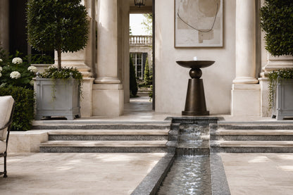 Stone pedestal fountain Kathib Ø65, pale warm stone, on a pale stone floor.