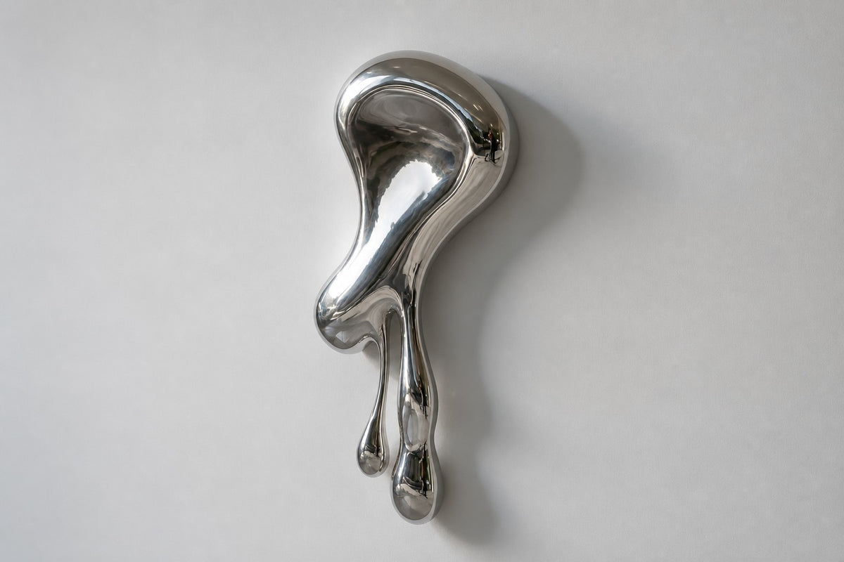 Lágrima — mirror-polished steel wall sculpture flowing down the wall into two drips, studio side angle showing its depth