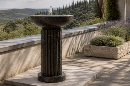Dark marble pedestal fountain Layla Ø65, deep black with white veining, on a pale stone floor.