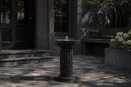 Dark marble pedestal fountain Layla Ø65, deep black with white veining, on a pale stone floor.