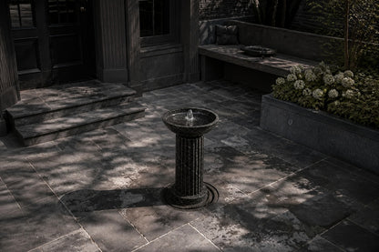 Dark marble pedestal fountain Layla Ø65, deep black with white veining, on a pale stone floor.