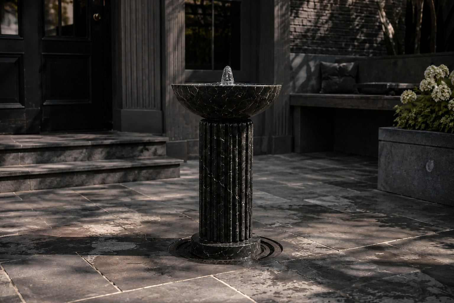Dark marble pedestal fountain Layla Ø65, deep black with white veining, on a pale stone floor.