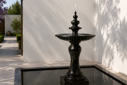 Stone pedestal fountain Nasima Ø65, black with gold and amber veining, on a pale stone floor.