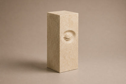 Tall cream Crema Marfil marble pedestal with a closed eye carved in shallow relief on one face.