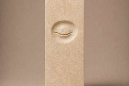 Tall cream Crema Marfil marble pedestal with a closed eye carved in shallow relief on one face.