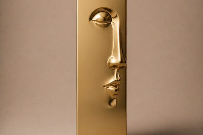Tall gold PVD pedestal with a closed eye, nose and mouth formed seamlessly out of one face.