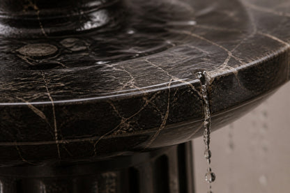 Stone pedestal fountain Sahran Ø65, black with gold veining, on a pale stone floor.