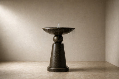 Dark marble pedestal fountain Sombra Ø65, dark olive-bronze, on a pale stone floor.