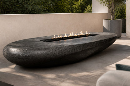 Charcoal-bronze stone fire table Suiran 220, charcoal with a warm bronze cast, on a pale stone floor.