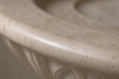 Pale limestone pedestal fountain Vasca Ø65, cream and white with amber veining, on a pale stone floor.