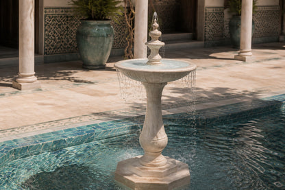 Pale limestone pedestal fountain Vasca Ø65, cream and white with amber veining, on a pale stone floor.